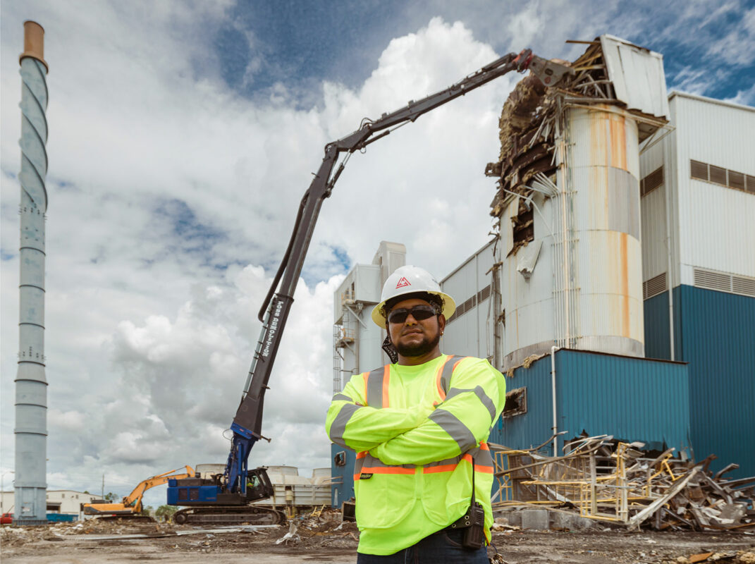 Man in safety vest and hard hat standing in front of building and high reach excavator